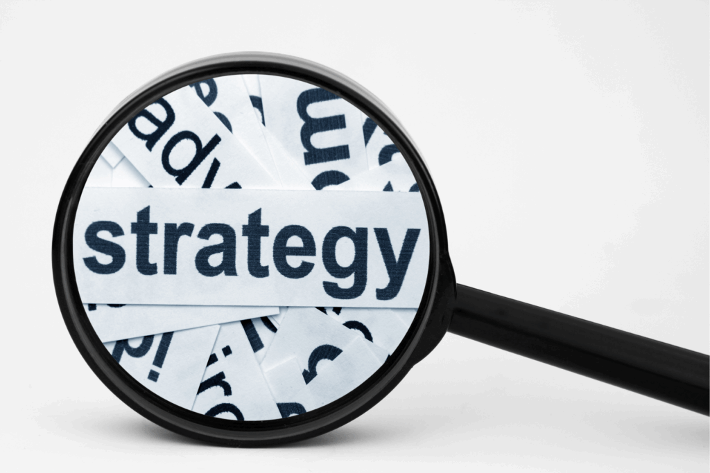 A magnifying glass inspects the word "strategy" typed on a thin strip of paper, lying atop many other such strips of paper.