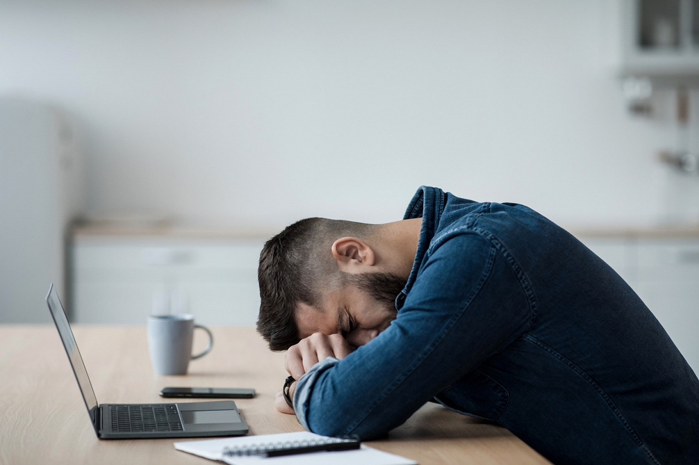A frustrated man sits at a desk with his head resting on his folded arms beside an open laptop, notebook, and coffee mug.